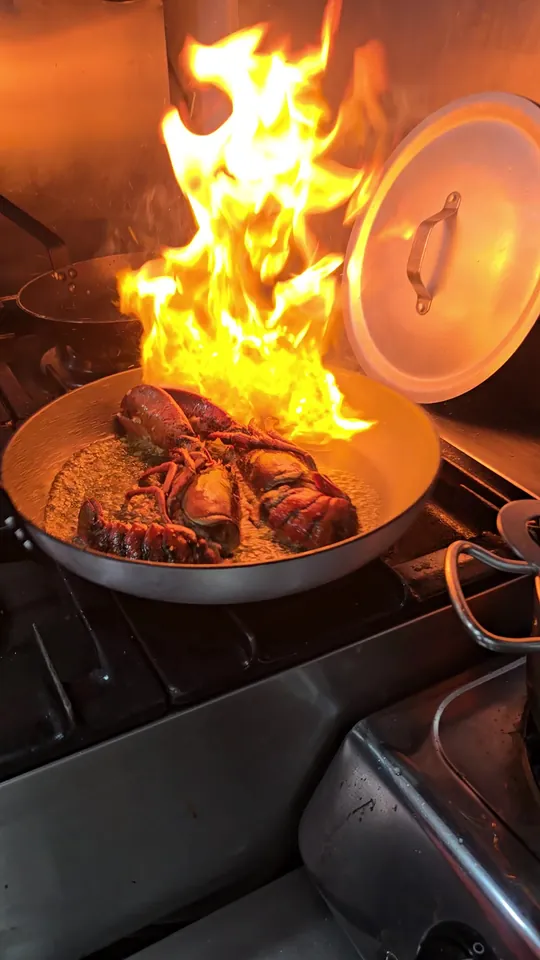 Una cola de bogavante flambeándose en una sartén sobre los fuegos de nuestra cocina: la llamarada sube alta y naranja desde la sartén, con la tapa apoyada a la derecha y otra sartén al fondo.