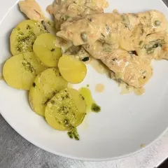 Un filete de pescado en salsa cremosa con hierbas picadas, junto a un abanico de patatas cocidas con aceite verde, en un plato blanco.