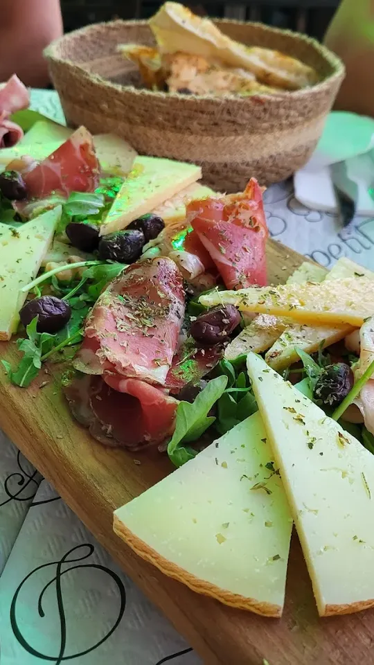 Una tabla de madera con cuñas de queso curado, lonchas de embutido, aceitunas negras, tomate y hojas de rúcula, y al fondo un cesto de pan.