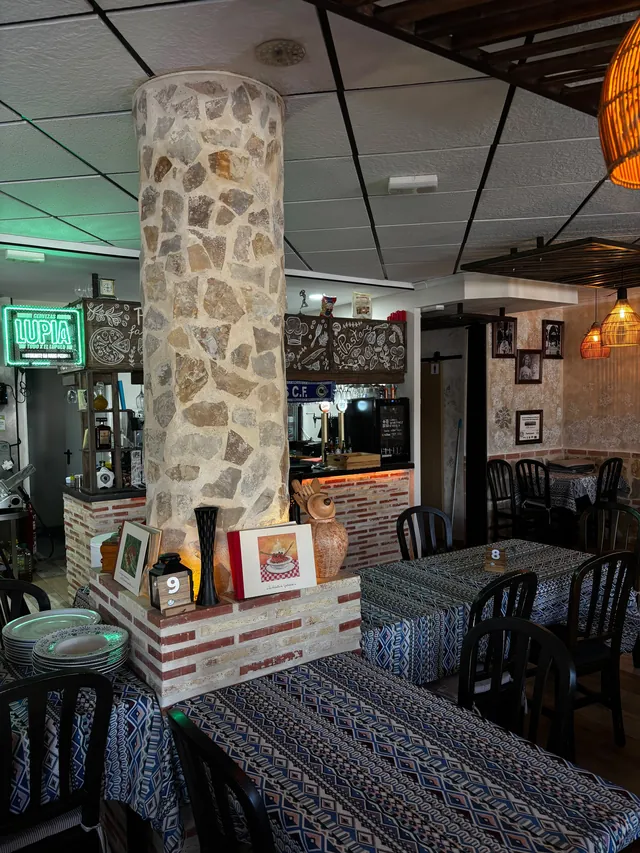 El comedor con la columna redonda forrada de piedra clara en el centro, sobre un zócalo de ladrillo donde hay un cuadro pequeño, un jarrón negro, una jarra de cerámica y el número 9 de una mesa. Alrededor, mesas con mantel estampado en azul y blanco, sillas oscuras y sus números; al fondo, la barra con los tiradores de cerveza, el rótulo de neón verde de Lupia y la pizarra dibujada, y a la derecha una pared con fotos enmarcadas y lámparas de mimbre.