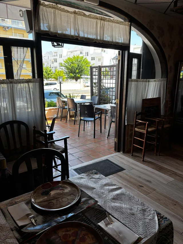 Desde dentro del comedor, mirando a la terraza por la puerta de cristal abierta: visillos blancos, el suelo de barro de la terraza con mesas blancas y sillas oscuras y, más allá, los árboles, los coches y los edificios blancos de la calle. En primer plano, una mesa puesta con mantel estampado, cubiertos, servilleta y dos bandejas oscuras, una de ellas con una pizza.