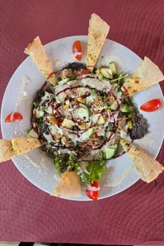 Una ensalada vista desde arriba: brotes, pepino, maíz, tomate cherry y queso rallado, con cuñas de pan plano puestas alrededor del plato.