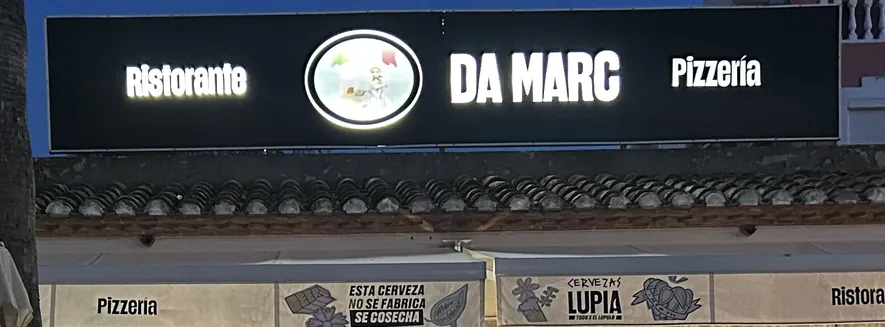 Nuestro rótulo encendido de noche sobre la fachada: sobre fondo negro, «Ristorante», el óvalo con el pizzero de nuestro logotipo, «DA MARC» en letras grandes y «Pizzería»; debajo, el alero de teja árabe y la banda del toldo, con «Pizzeria», «Ristorante» y dos anuncios de cerveza Lupia impresos en la lona.