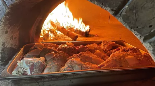 La boca de nuestro horno de leña con el fuego encendido y un tronco ardiendo dentro; delante, una bandeja metálica con piezas de carne cubiertas de queso y salsa de tomate.