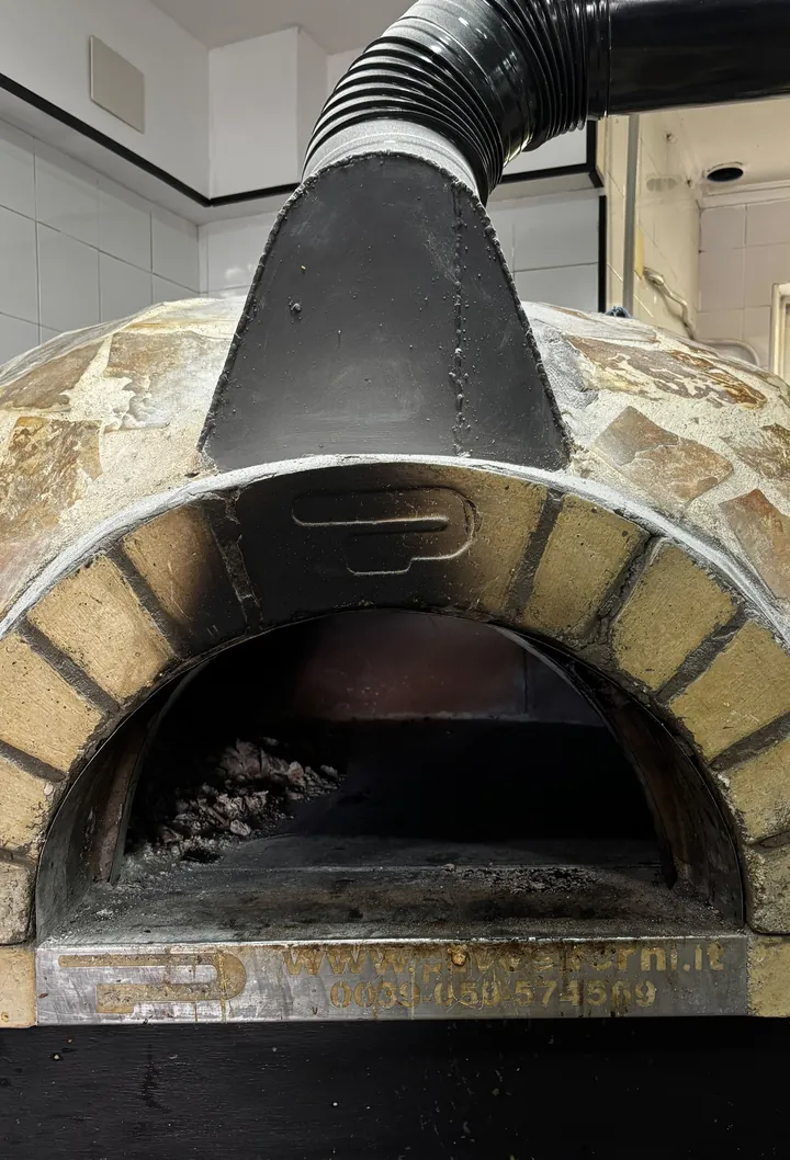 Nuestro horno de leña de frente, apagado: la cúpula forrada de piedra irregular, el arco de ladrillo amarillo alrededor de la boca y, dentro, la ceniza y la brasa del último servicio. Encima arranca el tubo negro de la chimenea; abajo, en el umbral, una chapa de latón grabada con la dirección web y el teléfono del fabricante del horno. Al fondo, la pared de azulejo blanco de la cocina.