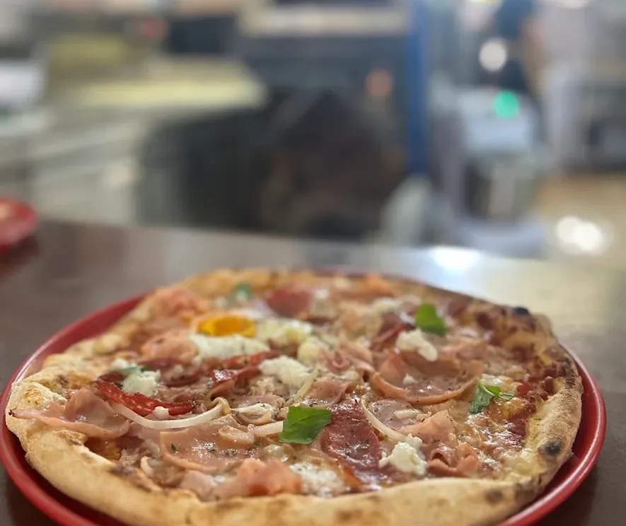 Una pizza recién hecha en un plato rojo, vista de cerca: jamón, salami, trocitos de queso fresco, hojas de albahaca y un huevo en el centro; detrás, desenfocado, el mostrador de la cocina.