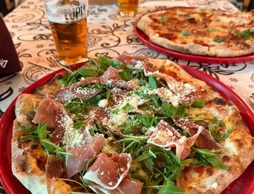 Una pizza con jamón, rúcula y queso rallado en un plato rojo, sobre un mantel estampado.