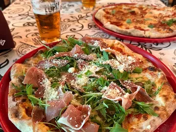 Una pizza con jamón, rúcula y queso rallado, en un plato rojo.