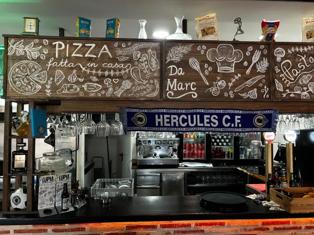 Nuestra barra: encima, tres tableros de madera oscura escritos con tiza — «PIZZA fatta in casa» con una pizza y los ingredientes dibujados, «Da Marc» bajo un gorro de cocinero, y «Pasta» —, y sobre ellos un reloj, paquetes de pasta y dos botellas blancas de cerámica. Debajo cuelgan las copas boca abajo y una bufanda azul del Hércules C.F.; en el mostrador negro, la cafetera, los tiradores de cerveza, las neveras de bebidas, botellas de cerveza Lupia con las cartas y una caja de naranjas.